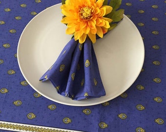 May include: A blue and yellow patterned napkin is folded and placed on a white plate. A yellow flower is used as a napkin ring.