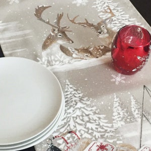 May include: A white table runner with a winter scene featuring deer and snowflakes. The runner is decorated with a red candle holder and two white plates.