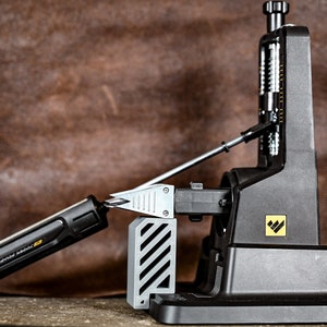 Może przedstawiać: Czarno-szary ostrzał do narzędzi z srebrnym ostrzem do ostrzenia. Czarno-szary śrubokręt jest ostrzony na ostrzu. Ostrzał do narzędzi ma żółte logo z trzema czarnymi trójkątami.