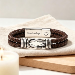 May include: A brown braided leather bracelet featuring a silver-toned metal clasp engraved with the text Never lose hope and a heart symbol. The bracelet includes a central silver-toned infinity-style knot detail with ribbed metal accents.
