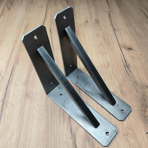 Puede incluir: Dos escuadras de estantería industriales de metal. Cada escuadra tiene una placa de montaje rectangular con tres agujeros y una barra de soporte diagonal. Las escuadras son de metal gris oscuro y están diseñadas para montaje en pared.