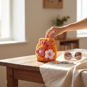 May include: A handmade orange crochet drawstring bag with two pink and white flower appliqués. The bag is sitting on a wooden table next to sunglasses. The bag has a pink drawstring closure.