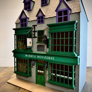 May include: A detailed model shop with a grey brick exterior, green window frames, and purple window surrounds. The sign above the shop reads "MAGICAL MENAGERIE". The roof features dormer windows with purple detailing. A sign for "FLEA POWDER" hangs from the building.