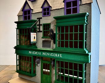 May include: A miniature model of a shop with a gray brick facade, green trim, and purple window frames. The sign reads "MAGICAL MENAGERIE" in white lettering. The roof has dormer windows with purple accents. A sign for "FLEA POWDER" hangs from the building.