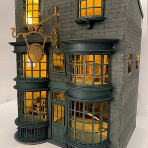 May include: A detailed model of a shop with a green door and large windows. The shop has a sign above the door with a circular design and the words "Ollivanders" written on it. The shop is made of gray brick and has a dark green roof.