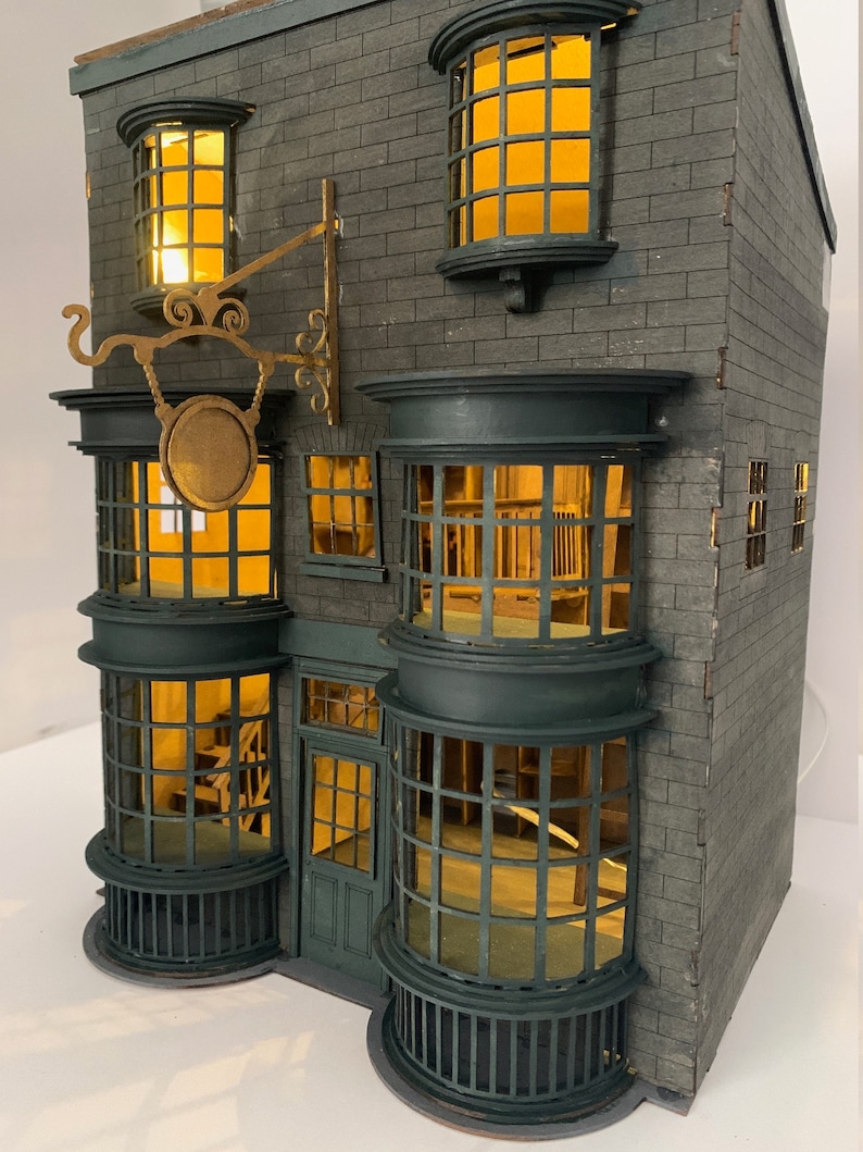 May include: A detailed model of a shop with a green door and large windows. The shop has a sign above the door with a circular design and the words "Ollivanders" written on it. The shop is made of gray brick and has a dark green roof.