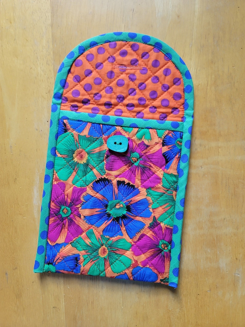Puede incluir: Un posavasos acolchado con estampado floral en tonos rosa, azul, verde y naranja. El posavasos tiene un borde azul turquesa con lunares blancos y un bot&oacute;n azul turquesa.