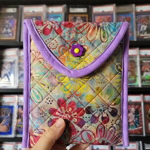 Puede incluir: Una funda de tel&eacute;fono acolchada con un patr&oacute;n floral en tonos de rosa, amarillo, azul y verde. La funda tiene un borde morado y un bot&oacute;n morado con una flor amarilla.