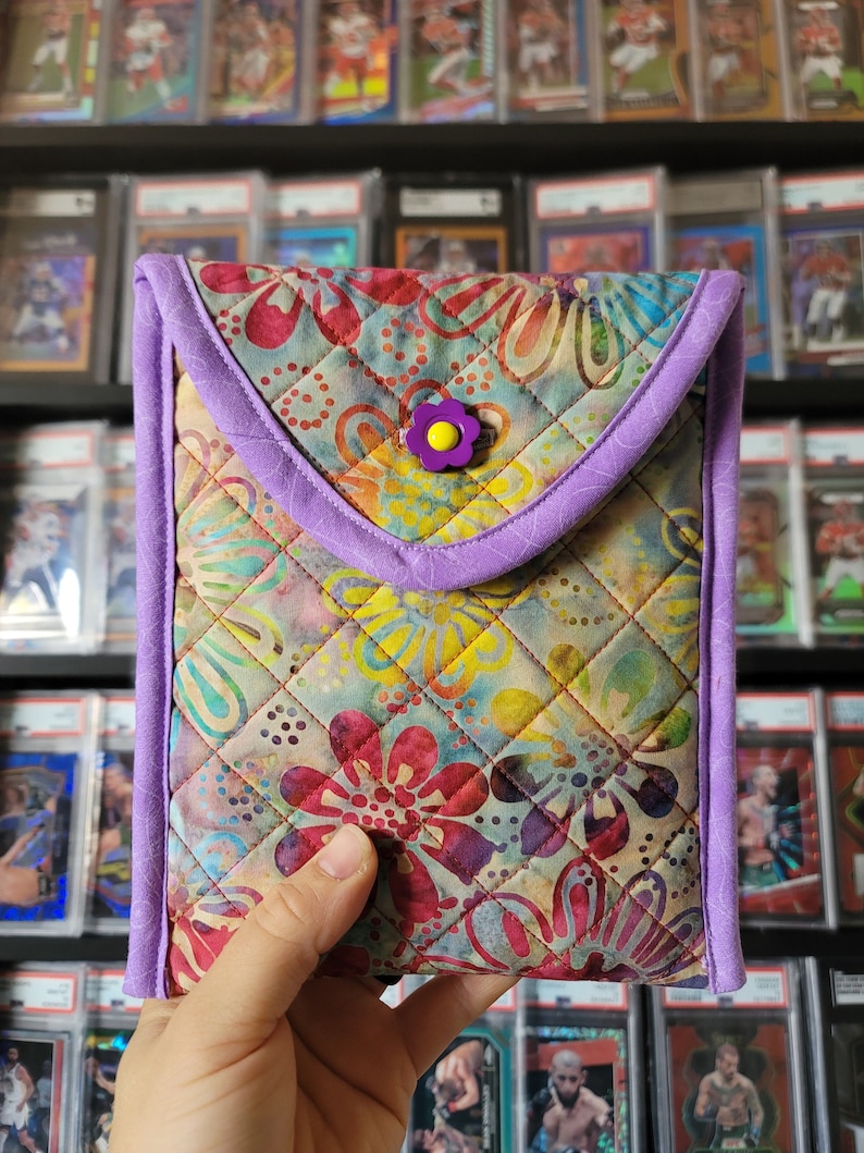 Puede incluir: Una funda de tel&eacute;fono acolchada con un patr&oacute;n floral en tonos de rosa, amarillo, azul y verde. La funda tiene un borde morado y un bot&oacute;n morado con una flor amarilla.