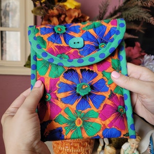 Puede incluir: Una funda para tel&eacute;fono floral de color naranja brillante, verde, azul y rosa con un bot&oacute;n turquesa y un ribete de lunares verdes y azules.