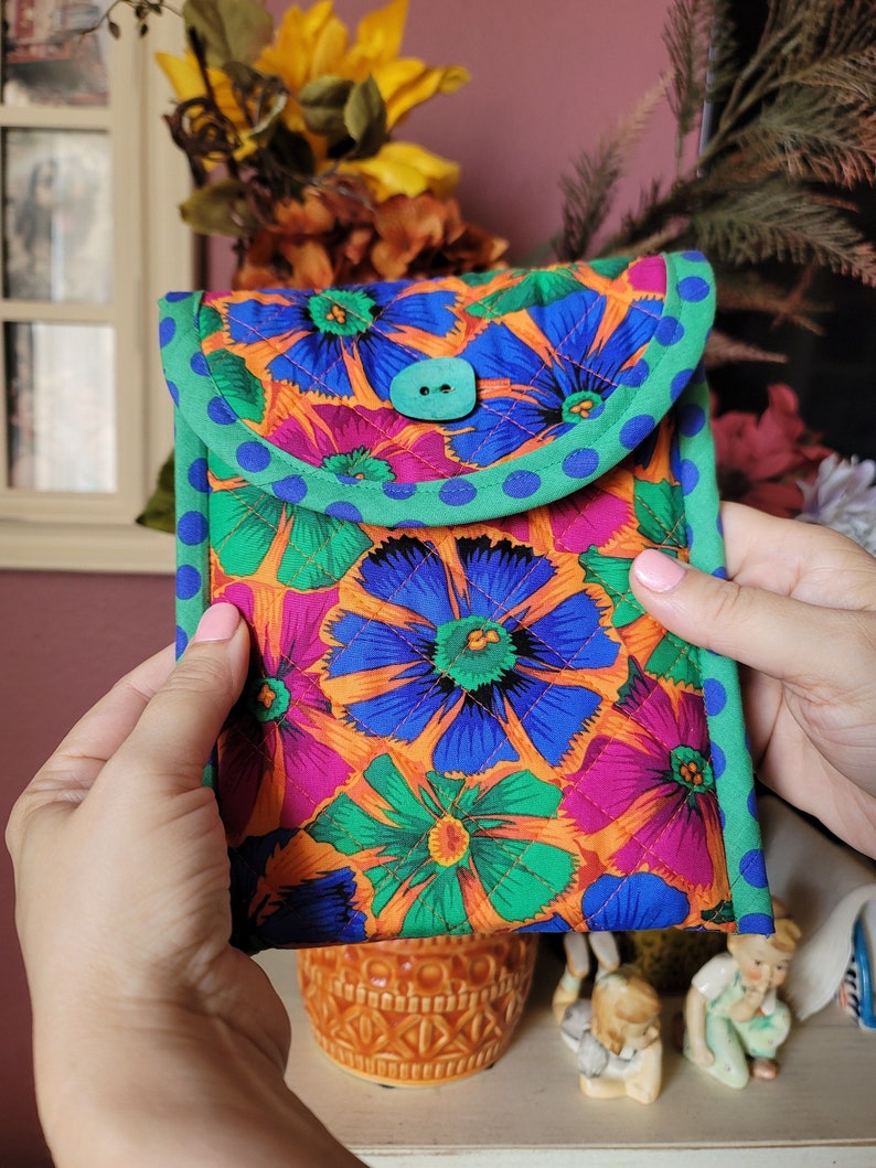 Puede incluir: Una funda para tel&eacute;fono floral de color naranja brillante, verde, azul y rosa con un bot&oacute;n turquesa y un ribete de lunares verdes y azules.