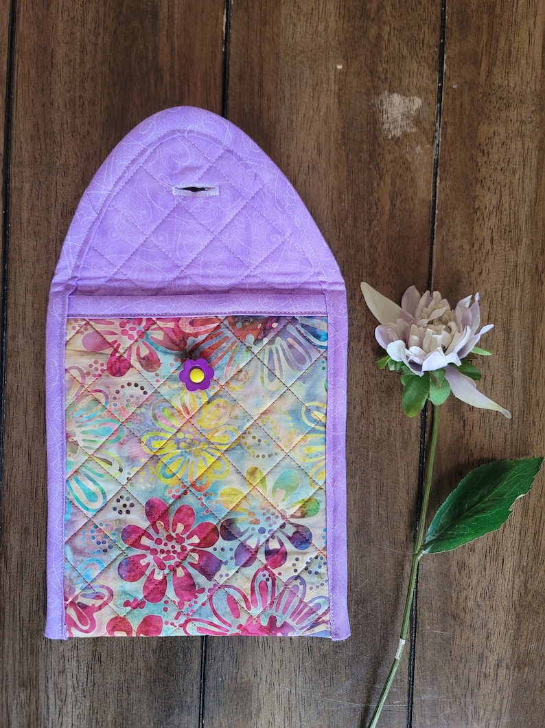 Puede incluir: Una bolsa acolchada de color morado con un forro de estampado floral y un cierre de bot&oacute;n morado. La bolsa est&aacute; sobre una superficie de madera.