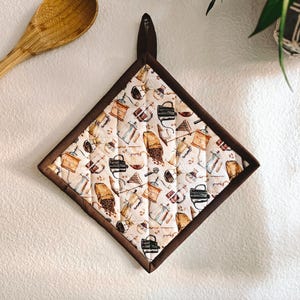 May include: A brown quilted pot holder with a coffee theme. The pot holder is decorated with a pattern of coffee beans, coffee pots, and other coffee-related items. The pot holder has a brown border and a loop for hanging.