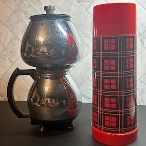 May include: A vintage chrome coffee pot with a black handle and base, next to a red and black plaid thermos. The coffee pot has a bulbous shape with a lid and a small knob. The thermos has a red top and bottom.