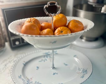 May include: A two-tiered serving dish filled with oranges. The white porcelain dish has a floral design with blue and pink accents and a silver trim. The oranges are bright orange and the dish is on a white countertop.