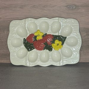 May include: A white ceramic egg holder with a rectangular shape and a basket-like design. The holder has 12 egg-shaped indentations and is decorated with red strawberries, yellow flowers, and green leaves. A decorative kitchen item.