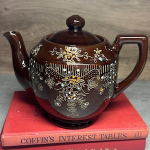 May include: A brown ceramic teapot with a lid, spout, and handle. The teapot is decorated with floral and geometric patterns in white, orange, and blue. It sits on top of two red books.