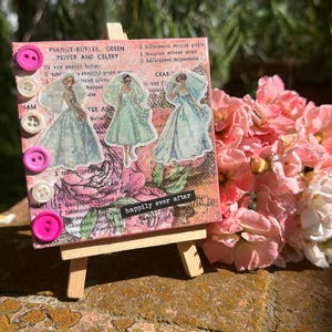 May include: A pink canvas with a floral pattern and a vintage recipe for peanut butter, green pepper and celery. The canvas is decorated with three brides in vintage wedding dresses and the words "happily ever after". The canvas is on a small wooden easel.