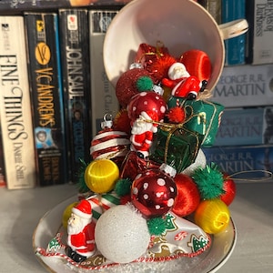 May include: A white teacup and saucer overflowing with Christmas ornaments. The ornaments include red, white, and gold baubles, miniature Santas, and small green gift boxes. The teacup is set against a backdrop of books.