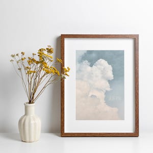 Peut inclure: Une impression encadrée représentant un paysage nuageux dans des tons bleus et beiges. L'impression est dans un cadre en bois marron avec un passe-partout blanc. Un vase avec des fleurs jaunes est à gauche. Le texte sur l'impression dit: "Les eaux calmes que l'âme était censée voir."