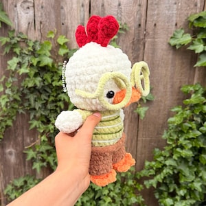 May include: A handmade crochet chicken doll with a white head, red comb, and yellow glasses. The chicken wears a green and white striped shirt, brown shorts, and orange feet. The doll is held in front of a wooden fence and green foliage.