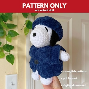 May include: A crocheted Snoopy doll wearing a blue coat and hat. The doll has a white body, black ears, and a black button on the coat. The text "PATTERN ONLY not actual doll" is at the top.