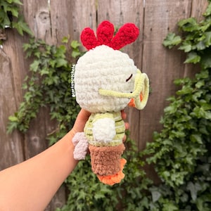 May include: A handmade crochet chicken plushie. The plushie has a white body, red comb, and yellow beak. It wears a green and white striped shirt and brown pants. The chicken's feet are orange. The plushie is held in front of a wooden fence.