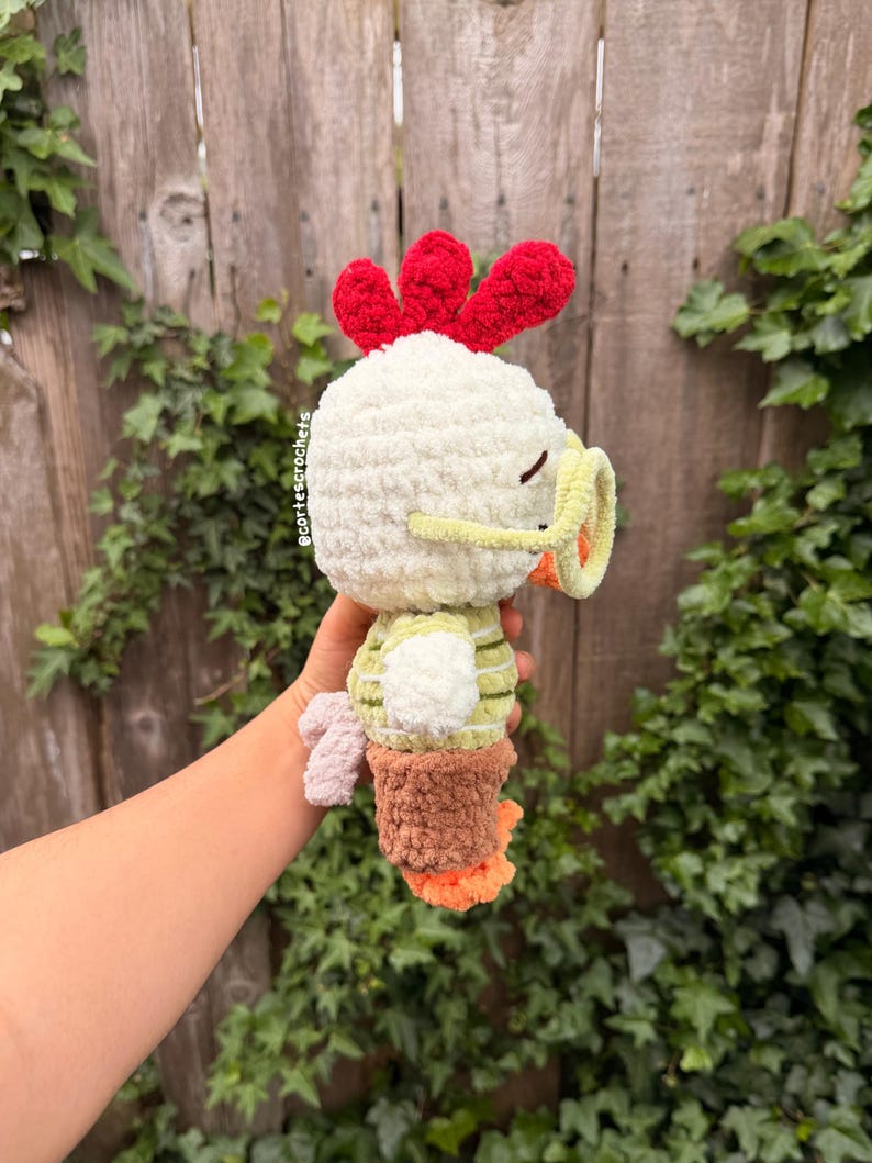 May include: A handmade crochet chicken plushie. The plushie has a white body, red comb, and yellow beak. It wears a green and white striped shirt and brown pants. The chicken's feet are orange. The plushie is held in front of a wooden fence.
