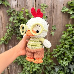 May include: A handmade crochet chicken toy with a white head, red comb, and yellow glasses. The chicken wears a green and white striped shirt, brown shorts, and orange feet. The toy is soft and plush, perfect for children.