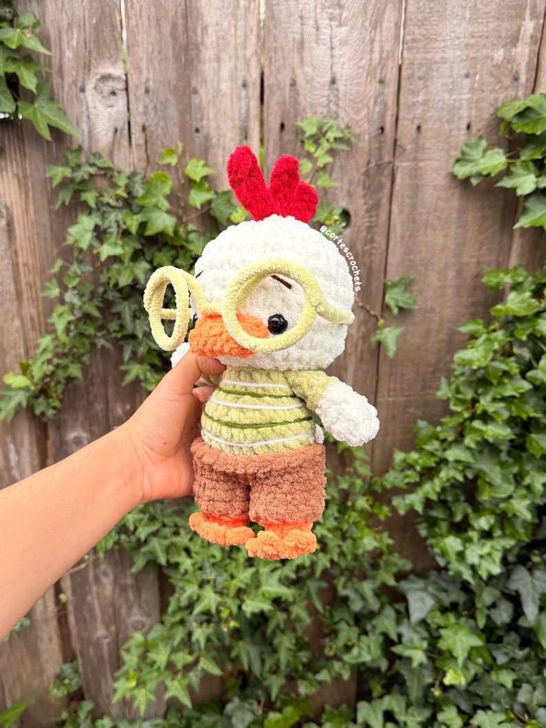 May include: A handmade crochet chicken toy with a white head, red comb, and yellow glasses. The chicken wears a green and white striped shirt, brown shorts, and orange feet. The toy is soft and plush, perfect for children.