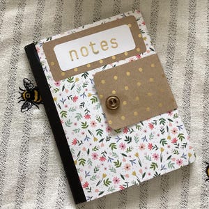 May include: A floral-patterned notebook with the word "notes" in gold lettering on the cover. The notebook has a brown flap with a button closure. The background is a striped fabric with embroidered bees.