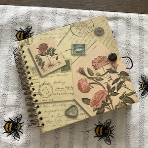 May include: A spiral-bound journal with a vintage aesthetic. The cover features floral illustrations, postage stamps, and handwritten script. A black button and a metal ring are attached to the cover. The journal is placed on a white and gray striped surface with embroidered bees.