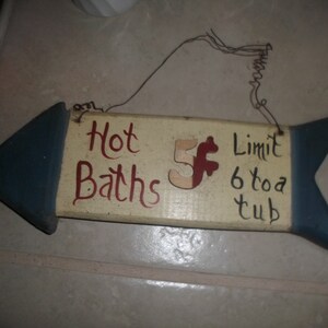 May include: A wooden sign with a blue arrow shape on each end. The sign reads "Hot Baths 5 Limit 6 to a tub".