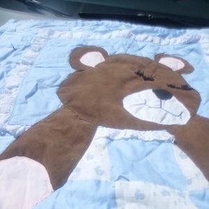 May include: A blue and white patchwork baby quilt with a brown teddy bear applique. The bear has closed eyes and a white smile.