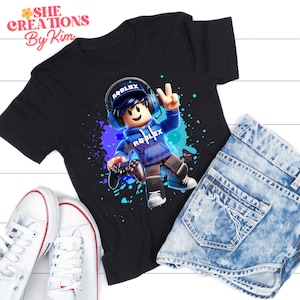 Może przedstawiać: Czarny T-shirt z grafiką postaci z kresk&oacute;wek w niebieskim swetrze i słuchawkach. Postać trzyma kontroler do gier wideo i ma napis "Roblox" na swetrze. Tło to niebieski i fioletowy wz&oacute;r rozprysku.