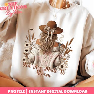 May include: White sweatshirt featuring an illustration of a person wearing a brown hat and holding a mug, surrounded by flowers and wheat. The text "You Are Made To Bloom" is below the illustration. The top left corner has the text "SHE CREATIONS By Kim".