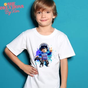 Może przedstawiać: Biała koszulka z grafiką niebieskiego i białego bohatera Roblox. Postać ma na sobie niebieską bluzę z kapturem, słuchawki i trzyma kontroler do gier wideo. Grafika jest otoczona niebieskimi i fioletowymi plamami farby.