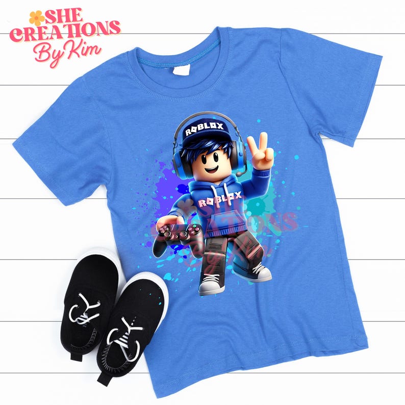 Może przedstawiać: Niebieska koszulka z grafiką postaci Roblox trzymającej kontroler do gier wideo. Postać ma na sobie niebieską bluzę z napisem "Roblox". Tło to akwarelowy wz&oacute;r w kolorze niebieskim i fioletowym.