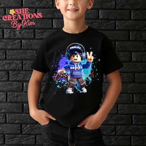 Może przedstawiać: Czarny T-shirt z kolorową grafiką postaci Roblox trzymającej kontroler do gier wideo. Postać ma na sobie niebieską bluzę z kapturem i słuchawki. Grafika jest otoczona niebieskimi i fioletowymi plamami farby.