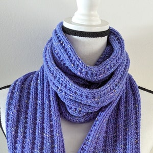 May include: A purple knitted scarf with a speckled pattern. The scarf is folded and draped over a white mannequin.