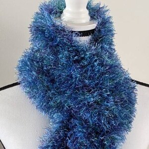 May include: A long, textured scarf made of fuzzy, eyelash-style yarn in various shades of blue, including deep navy, bright royal blue, and hints of teal. The material has a shaggy, multi-dimensional appearance with a soft, voluminous pile.