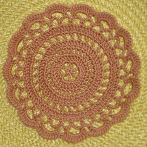 May include: A close-up of a crocheted doily with a brown center and a light brown outer edge. The doily is on a yellow woven surface.