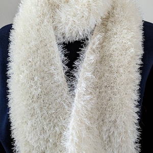 May include: Cream-colored, fluffy infinity scarf. The scarf is made of a soft, textured material, creating a cozy and warm appearance. The scarf is draped around a mannequin's neck, showcasing its full, circular design.