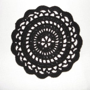 May include: A black, circular, crocheted doily with a scalloped edge. The doily features concentric circles of openwork, creating a delicate and intricate design. The handmade piece is set against a white background.