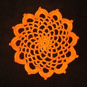 May include: An orange, crocheted doily with a floral design. The doily is circular with a central spiral pattern and a scalloped edge. The openwork design allows the black background to show through.