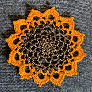 Könnte beinhalten: Eine handgefertigte, gehäkelte Tischdecke mit floralem Design. Die Mitte ist dunkelbraun, umgeben von einem Ring aus Lochmuster und mit einem hellorangen, gewellten Rand versehen. Die Tischdecke ist kreisförmig und liegt auf einer blauen Denim-Oberfläche.