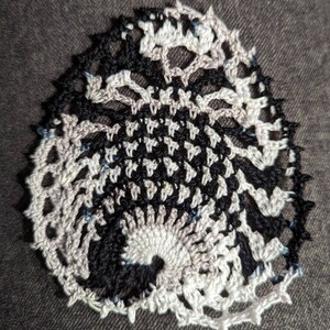 May include: A black and white crocheted doily with a spiral design. The doily is made with a fine yarn and has a delicate, lacy appearance.