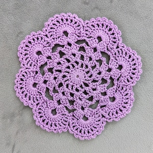 May include: A handmade, lavender-colored crochet doily with a floral design. The doily features a central spiral pattern surrounded by openwork and scalloped edges. The intricate details and soft color make it a decorative item.
