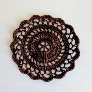 May include: A handmade, brown crocheted doily with a circular design. The doily features a detailed openwork pattern with a scalloped edge. The intricate design is centered with a spiral pattern, creating a decorative and delicate piece.
