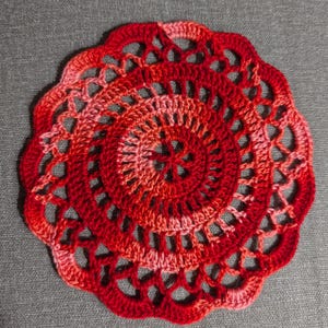May include: A red and pink crocheted doily with a circular design. The doily has a delicate lace pattern and is made with a soft yarn.
